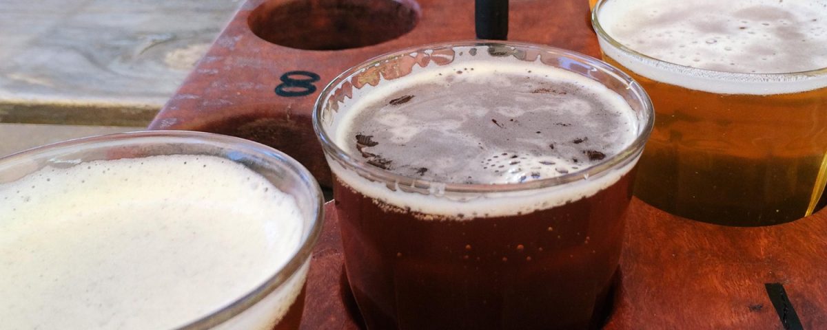 Bandeja com amostras de cervejas artesanais de diferentes cores, destacando o processo de carbonatação de bebidas artesanais. A imagem mostra uma bandeja de degustação com seis copos de cervejas artesanais, variando de tons âmbar a marrom escuro, que apresentam níveis variados de carbonatação e colarinho. A disposição das bebidas em diferentes cores e texturas sugere uma variedade de estilos artesanais, cada um com um nível específico de gás carbônico, essencial para realçar o sabor e a experiência sensorial.