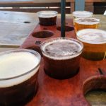 Bandeja com amostras de cervejas artesanais de diferentes cores, destacando o processo de carbonatação de bebidas artesanais. A imagem mostra uma bandeja de degustação com seis copos de cervejas artesanais, variando de tons âmbar a marrom escuro, que apresentam níveis variados de carbonatação e colarinho. A disposição das bebidas em diferentes cores e texturas sugere uma variedade de estilos artesanais, cada um com um nível específico de gás carbônico, essencial para realçar o sabor e a experiência sensorial.