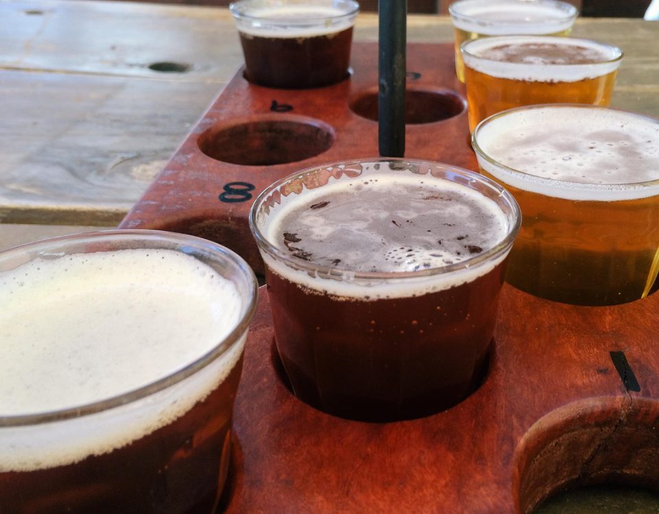 Bandeja com amostras de cervejas artesanais de diferentes cores, destacando o processo de carbonatação de bebidas artesanais. A imagem mostra uma bandeja de degustação com seis copos de cervejas artesanais, variando de tons âmbar a marrom escuro, que apresentam níveis variados de carbonatação e colarinho. A disposição das bebidas em diferentes cores e texturas sugere uma variedade de estilos artesanais, cada um com um nível específico de gás carbônico, essencial para realçar o sabor e a experiência sensorial.