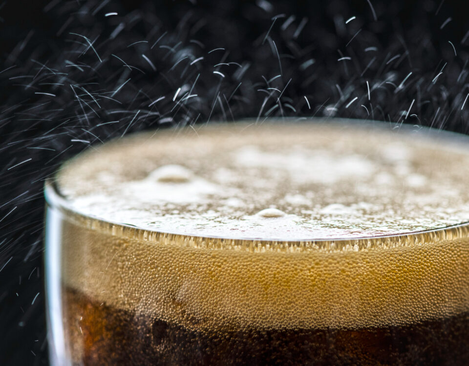 Imagem de um copo com líquido escuro e espuma na superfície, indicando um refrigerante ou bebida carbonatada. Pequenas bolhas são visíveis na espuma, enquanto gotículas e partículas de gás sobem do líquido, ilustrando o processo de carbonatação. A cena tem um fundo escuro que destaca o brilho das bolhas e a efervescência característica da carbonatação.