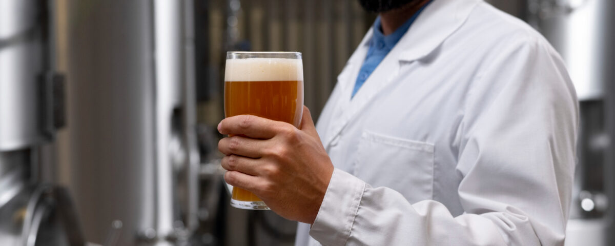 Homem usando jaleco branco segura um copo de cerveja artesanal, com coloração âmbar e uma camada de espuma cremosa no topo, em um ambiente industrial. Ao fundo, tanques de aço inoxidável, comuns em cervejarias, indicam o processo de produção e carbonatação de cerveja. A cena destaca a análise visual da bebida, um passo importante no controle de qualidade da carbonatação e da textura da espuma.