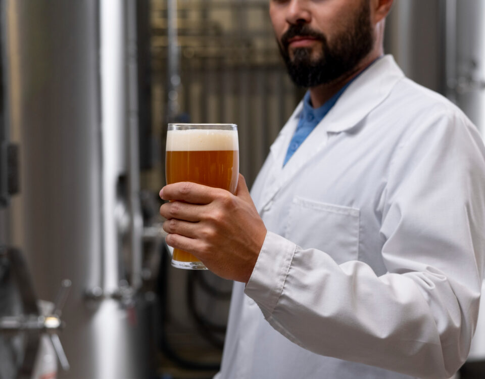 Homem usando jaleco branco segura um copo de cerveja artesanal, com coloração âmbar e uma camada de espuma cremosa no topo, em um ambiente industrial. Ao fundo, tanques de aço inoxidável, comuns em cervejarias, indicam o processo de produção e carbonatação de cerveja. A cena destaca a análise visual da bebida, um passo importante no controle de qualidade da carbonatação e da textura da espuma.